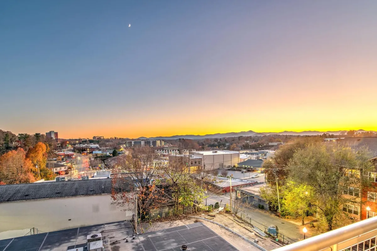 A stunning view of the Asheville cityscape at sunset, with the distant Blue Ridge Mountains and a colorful sky creating a dramatic backdrop. The autumn trees in the foreground add warmth and seasonal vibrancy. The image captures the ambiance of downtown Asheville as evening approaches, hinting at the vibrant nightlife and lively city atmosphere.