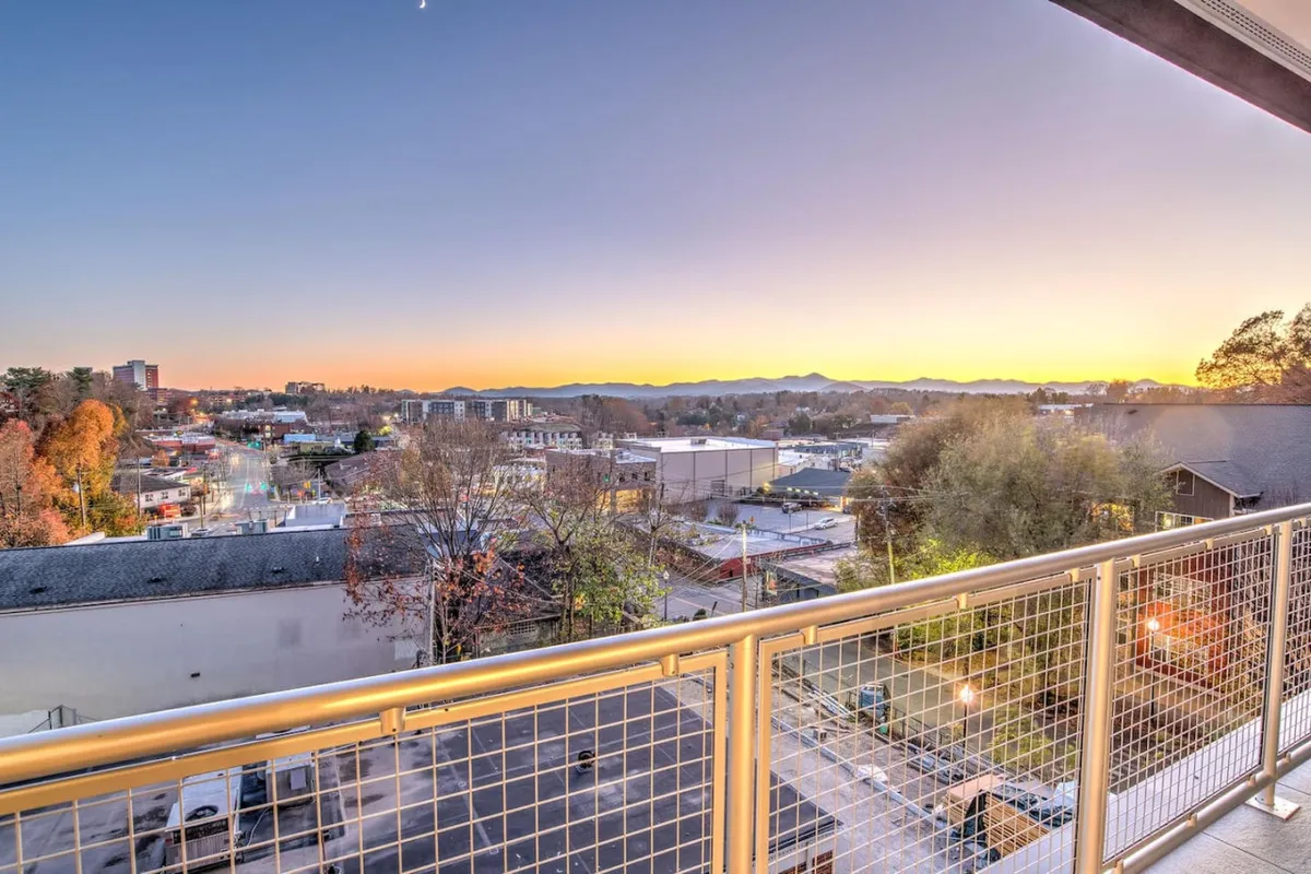 This view-focused image features a balcony railing framing the dramatic city skyline with the Blue Ridge Mountains rising in the distance. Sunset colors fill the sky, while the perspective situates the viewer as if experiencing the tranquil scenery firsthand from the balcony.
