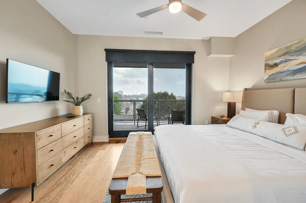 A serene bedroom setup with a large bed, wooden dresser, TV, and a sliding door opening to a balcony with views of the city. The room is bathed in soft light, hinting at the comfort and relaxation awaiting after a night out. The direct connection to the balcony emphasizes both indoor comfort and the ability to appreciate Asheville’s skyline from the privacy of the condo.