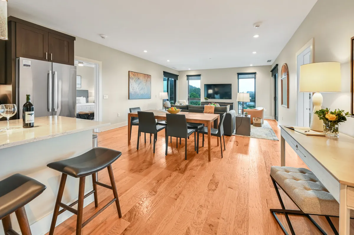 An inviting open-plan living area featuring a comfortable sofa, a modern dining table, and a large kitchen island. Floor-to-ceiling windows bathe the space in natural light and offer striking urban views. The space is styled for relaxation and social gatherings, with ample room for dining, entertaining, or unwinding with a favorite show.