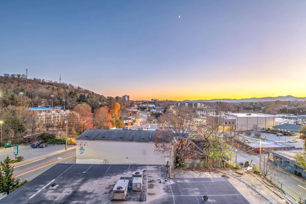 This image captures a stunning sunset over the Blue Ridge Mountains, with the Asheville city skyline and colorful autumn trees in the foreground. The view is expansive, evoking the romance and natural beauty central to the section theme.