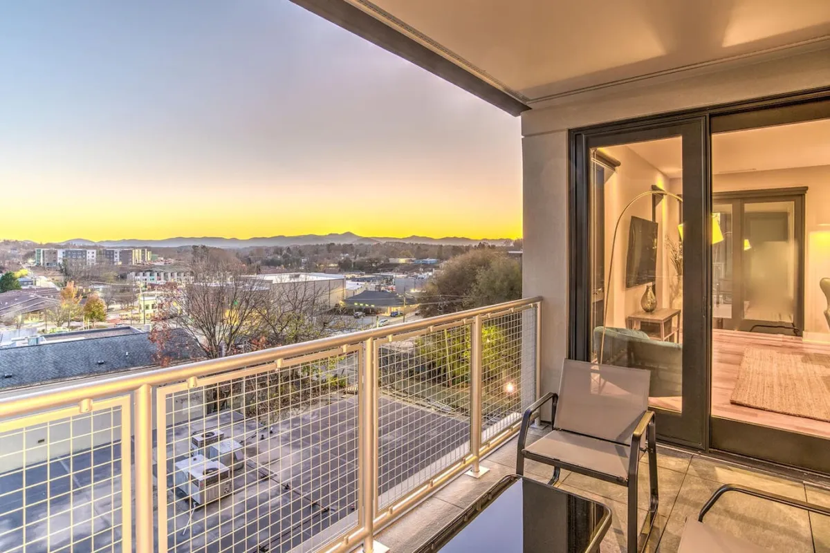An exterior shot of a balcony at dusk, showing the interplay of soft, ambient lighting from inside the suite and the glowing city lights beyond. The balcony railing and seating are visible, suggesting a perfect spot for evening relaxation or lively conversation while the sunset unfolds.