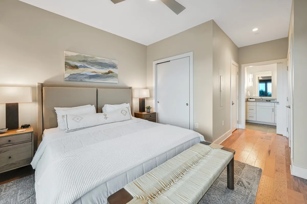 A serene king bedroom featuring a plush bed with luxury bedding, flanked by nightstands and gentle lighting. Artwork adorns the walls, and a doorway leads to a private ensuite bathroom. Soft morning light fills the room, reinforcing a tranquil, restful atmosphere.