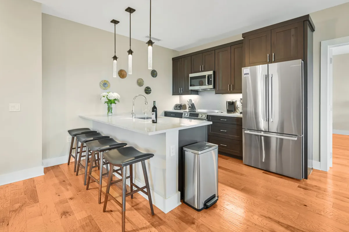 A modern kitchen with a large island, sleek appliances, bar stools, and decorative touches like fresh flowers and plates. The open flow into the living and dining spaces supports both everyday cooking and entertaining guests, echoing the condo’s focus on effortless living.