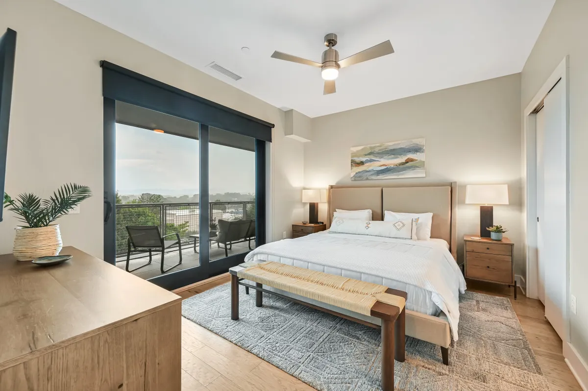 A serene bedroom with a comfortable bed facing large windows, nightstands with lamps, stylish artwork, and a bench at the foot of the bed. The window frames a scenic view, hinting at the natural beauty just beyond, while soft lighting creates a welcoming atmosphere.