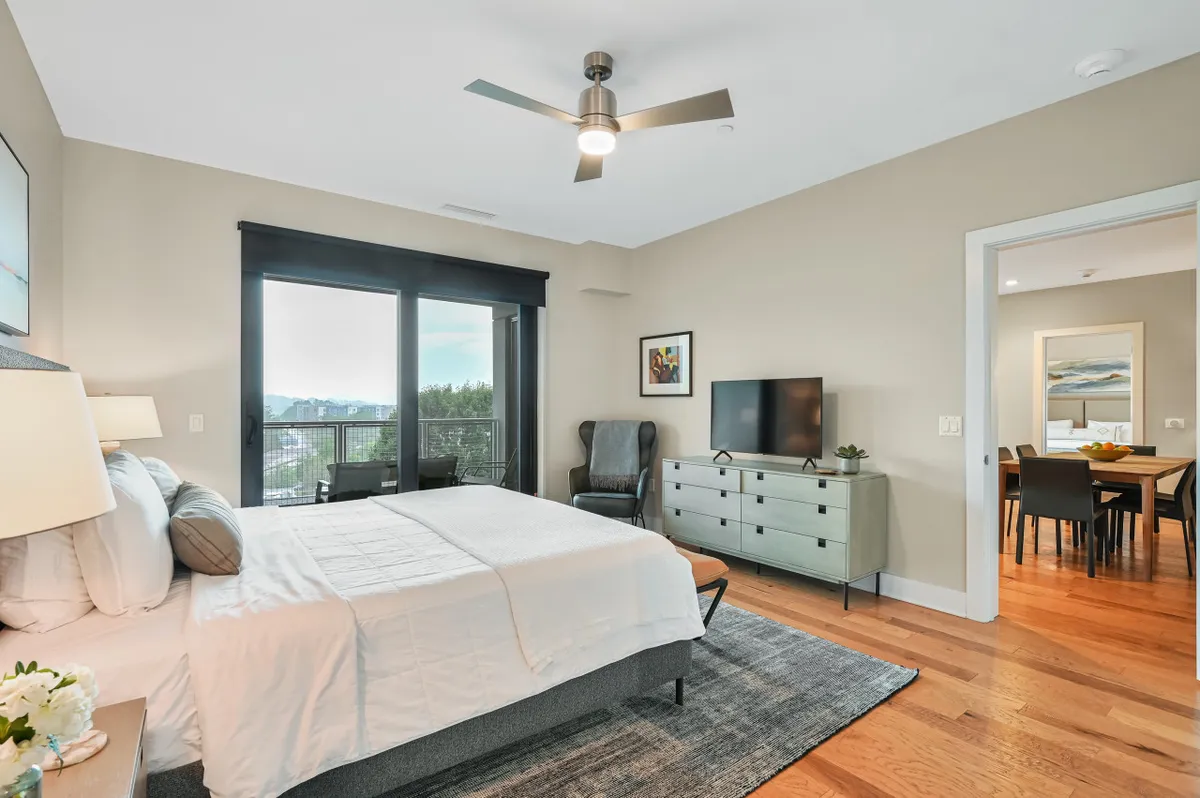 A calm and cozy bedroom featuring a plush bed, large windows with abundant natural light, an armchair, and a TV. There’s also a small dining area visible in the background, emphasizing multi-purpose use and comfort.