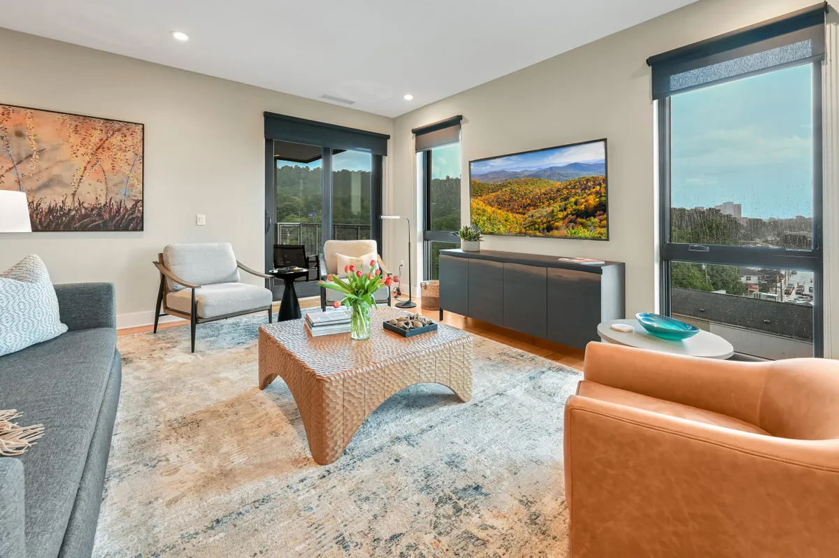 A stylish, inviting living room with a plush sofa, modern coffee table, and large windows framing sweeping views of the Blue Ridge Mountains. The space is bathed in natural light, perfect for relaxing, entertaining, or taking in breathtaking sunsets right from the comfort of your condo.
